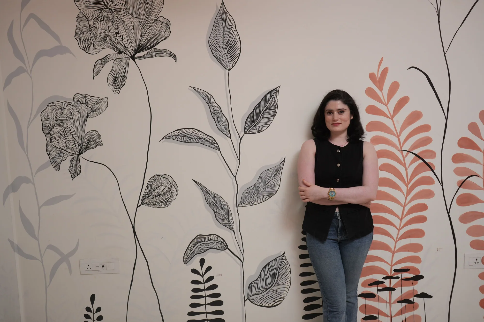 Yuvasri standing against a wall she painted with large ink botanicals and coral fronds