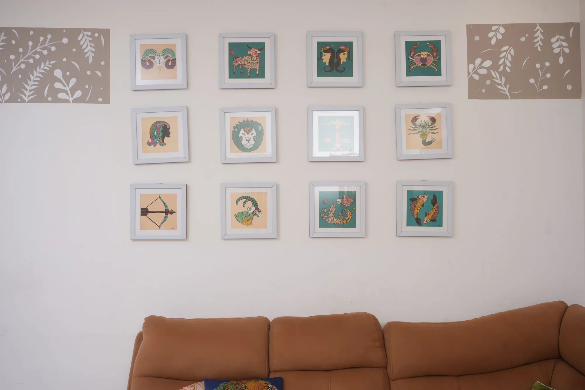 The twelve framed zodiac paintings hung together as a four-by-three grid on the wall above a sofa