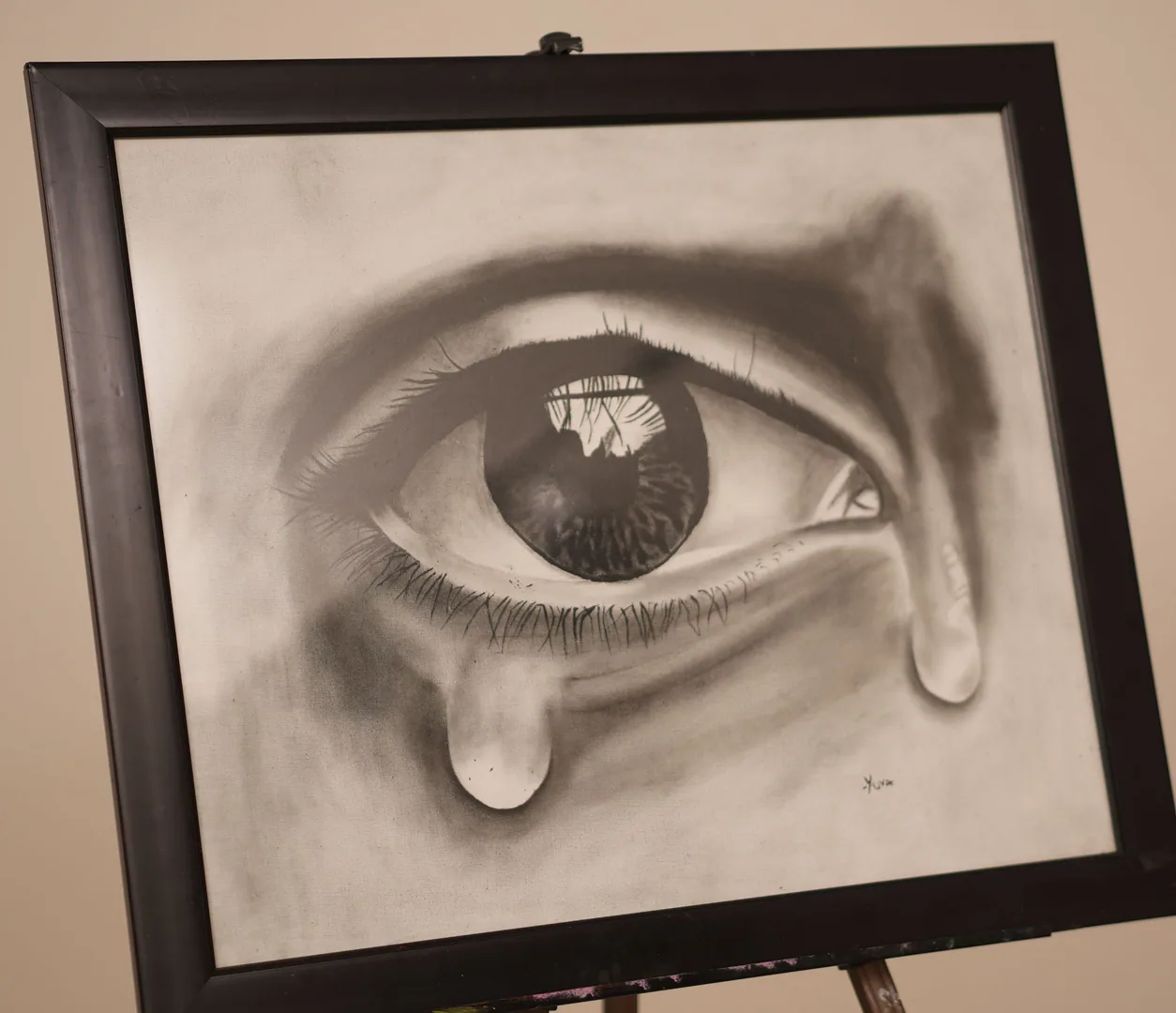 Large charcoal drawing of a single human eye with a tear falling from it, the iris carrying a reflection, framed in dark wood on an easel